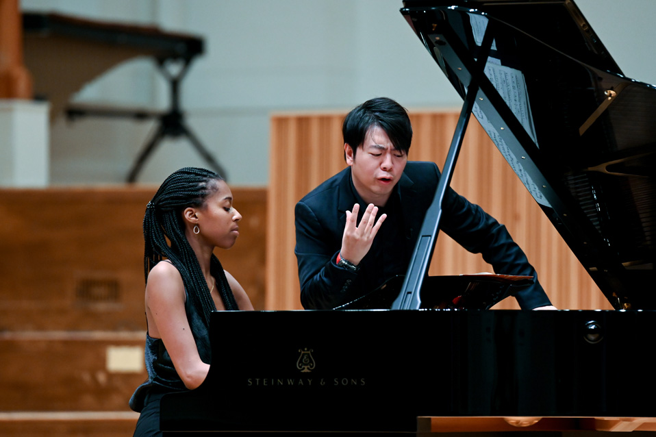 A black female student, performing on a piano, with Lang Lang, wearing a dark suit, guiding the student's performance.